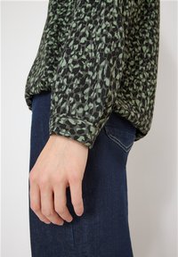 Hand resting by side wearing dark blue jeans and green blouse with black and light green animal print pattern.