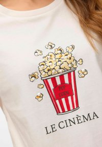 Biała koszulka z grafiką pudełka na popcorn w paski, z żółtym popcornem i dekoracyjnymi perłami; napis brzmi "LE CINÉMA."