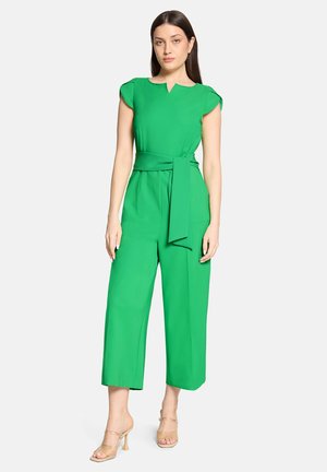 Femme aux longs cheveux foncés portant une combinaison verte vif à manches courtes, ceinture à la taille, et sandales à talons hauts beige, debout sur fond blanc.