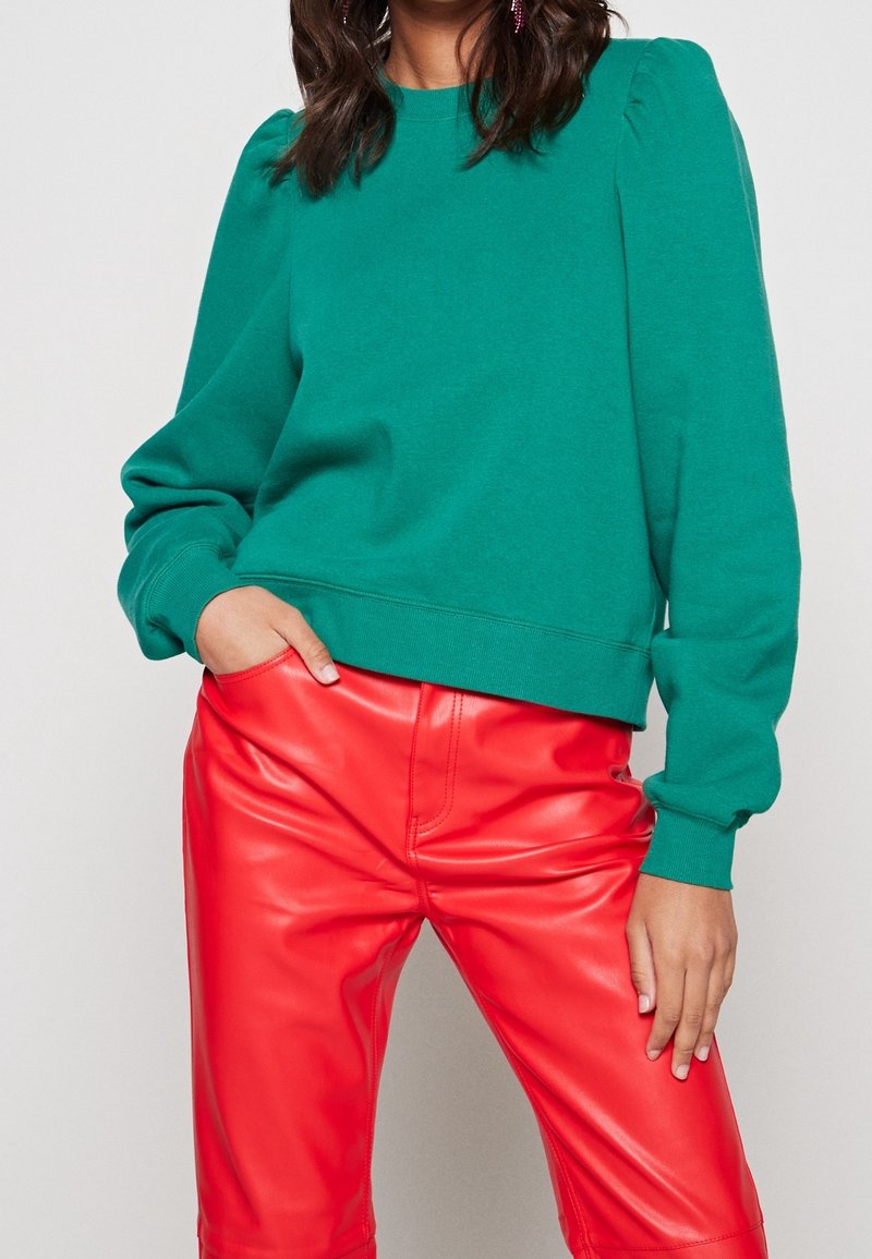 Femme portant un pull vert à manches longues avec des épaules bouffantes et un pantalon en cuir rouge vif, une main dans la poche et l'autre reposant sur la cuisse.