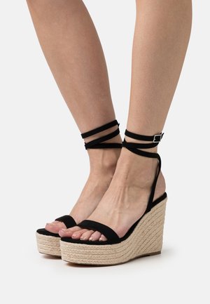 Las sandalias de cuña de ante negro cuentan con un tacón de yute trenzado y correas en los tobillos. El diseño incluye punta abierta y un acabado suave y texturizado.
