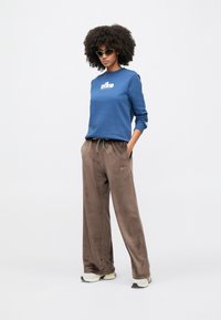 Άτομο με σγουρά μαλλιά που φοράει γυαλιά ηλίου, μπλε φούτερ, καφέ παντελόνι από βελούδο και λευκά αθλητικά παπούτσια, στέκεται μπροστά σε απλό φόντο.