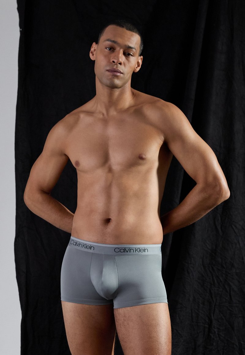 Graue Baumwoll-Boxershorts mit einer weichen, glatten Textur und einem elastischen Taillenbund, der das "Calvin Klein"-Logo in Schwarz zeigt. Das Modell steht mit entspannten Armen.