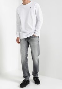 Camiseta blanca de manga larga con un pequeño logo, combinada con unos pantalones vaqueros grises desgastados y zapatos negros de cordones. El conjunto tiene un diseño casual.