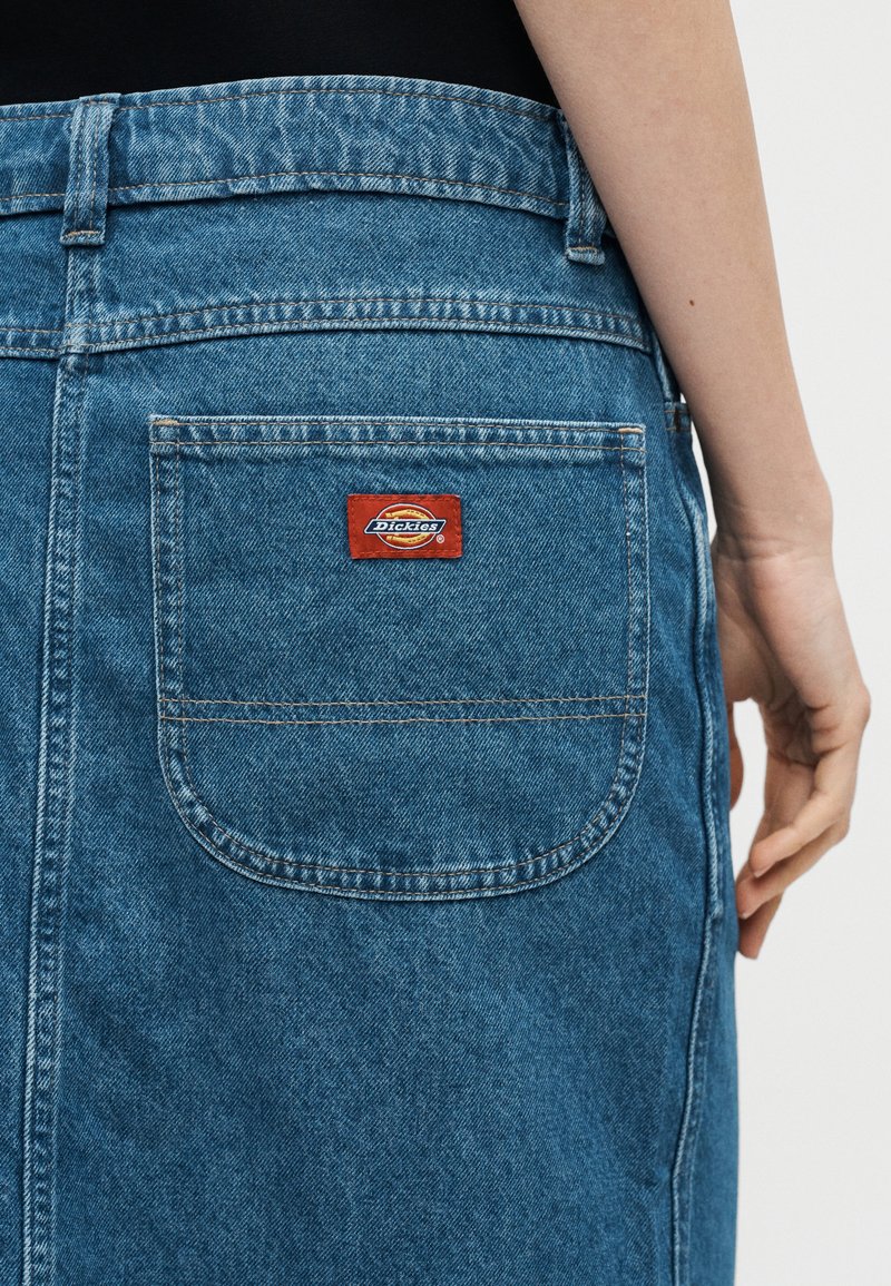 Rückenansicht einer Person, die einen blauen Jeansrock mit einer quadratischen Tasche auf der Rückseite trägt, die ein rotes Dickies-Logo-Patch zeigt.