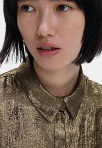 Camisa metálica dorada con cuello, con una textura brillante y sutiles rayas oscuras. Tiene un botón marrón en el cuello y en la tapeta de botones.