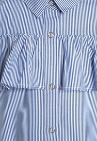 Niebiesko-biała koszula w paski z zapięciem na guziki, falbaniastym wzorem na piersi oraz srebrnymi pierścieniami na guzikach.