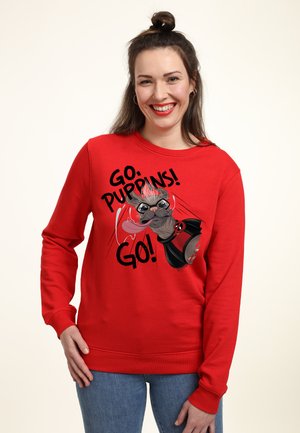 Femme souriante aux cheveux foncés en chignon porte un sweat-shirt rouge avec un chien de dessin animé et le texte "Go Puppies! Go!"