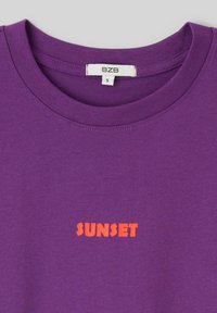 T-shirt violet à col rond avec le texte orange "SUNSET" centré sous le col et une étiquette de marque "BZB" à l'intérieur du col.