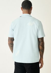 Camisa de manga corta en azul claro, hecha de una tela suave, con bordado de hojas blancas en la parte posterior y el lado izquierdo, con un ajuste relajado y cuello plano.