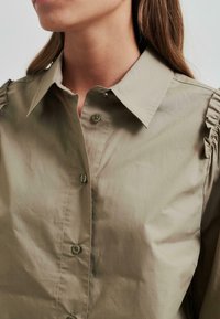 Šviesiai alyvuogių spalvos marškiniai, su sagomis, turi smailią apykaklę. Yra raukšlių detalės ant pečių, švelni tekstūra ir atitinkamos sagos.