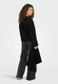Sort oversized jakke med firkantet snit og lange ærmer, parret med mørkegrå bukser med bred ben; model holder et matchende sort stof.