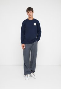 Felpa blu navy con scollo a crew e etichetta a patch, abbinata a pantaloni grigi a gamba larga e sneakers bianche. Sfondo neutro.