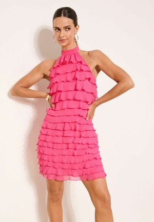 Pruunidega naine kõrge kaelusega varrukateta roosas volangiga kleidis, seistes käed puusadel heledal taustal.