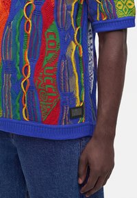 Gros plan d'une personne portant un haut court en tricot coloré avec des motifs bleus, rouges, verts et jaunes sur un jean bleu, montrant la main et le bras.