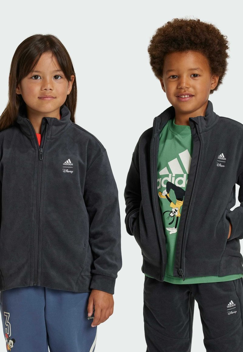 Giacche in pile grigie con zip frontale, con logo Adidas e marchio Disney. Accompagnate da una maglietta verde e pantaloni blu con design.