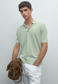 Polo a maniche corte in verde menta, in tessuto a maglia. Presenta una chiusura a tre bottoni e polsini a coste. Abbinato a pantaloni bianchi e borsa in camoscio marrone.