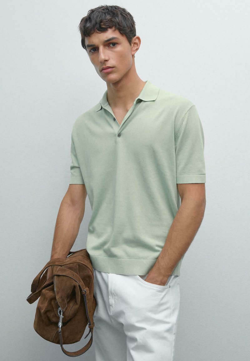 Polo a maniche corte in verde menta, in tessuto a maglia. Presenta una chiusura a tre bottoni e polsini a coste. Abbinato a pantaloni bianchi e borsa in camoscio marrone.