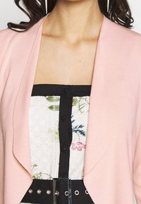 Cardigan rosa chiaro con apertura frontale, indossato sopra una maglietta a fantasia floreale bianca e nera. Presenta una cintura nera con borchie argentate in vita.