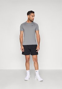 Szürke, rövid ujjú sportpóló fekete Nike logóval, fekete rövidnadrággal és fehér sportcipővel egy egyszínű háttéren.