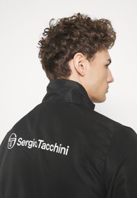 Fekete dzseki magas gallérral, texturált anyagból, fehér logónyomattal a hátsó részen. A dizájn stílusos és kortárs.