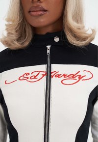 Nærbillede af person iført en sort og hvid lynlåsjakke med rød "Ed Hardy" broderi over brystet og en knappet krave.