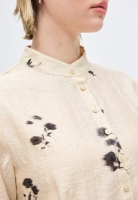 Γυναίκα που φοράει μια κρεμ μπλούζα με κουμπιά και μανδαρινέ γιακά, με μαύρα ανθισμένα σχέδια, εμφανιζόμενη από τον λαιμό μέχρι το στήθος.