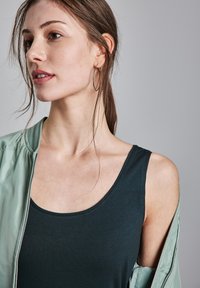 Canotta verde scuro con scollatura ampia, abbinata a una giacca bomber verde chiaro. La giacca ha una texture lucida e un colletto a coste.