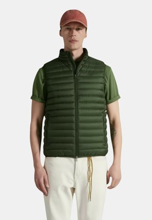 Uomo con maglietta verde a maniche corte, gilet imbottito verde scuro, pantaloni bianchi e cappellino da baseball rosso, in piedi davanti a uno sfondo uniforme.