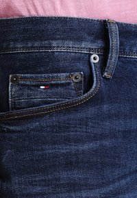 Mörkblå jeans i denim med en liten framficka, silvernitar och synliga gula sömmar. Ett rosa tyg skymtar delvis ovanför midjebandet.