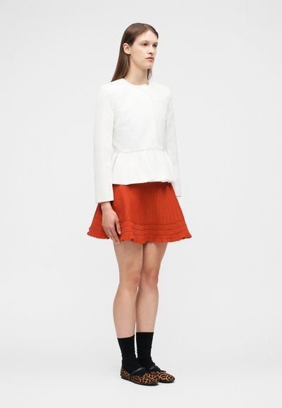 Νεαρή γυναίκα που φοράει λευκό μπλουζάκι με μακριά μανίκια, πορτοκαλί πλισέ φούστα, μαύρες κάλτσες και επίπεδα παπούτσια με λεοπάρ μοτίβο, στέκεται μπροστά σε λευκό φόντο.