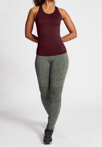Donna in piedi con le mani dietro la schiena, indossa un top senza maniche bordeaux, leggings verdi con texture e scarpe da ginnastica nere con dettagli gialli.