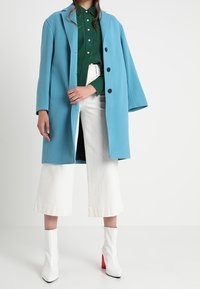 Cappotto di lana blu con un design oversize, dotato di bottoni neri, indossato sopra una camicia verde, abbinato a pantaloni bianchi a gamba larga e stivali bianchi.