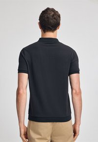 Svart polotröja med korta ärmar, texturerad tyg, ribbad krage och muddar, samt en figursydd nederkant. Inga synliga logotyper eller mönster.