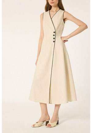 Femme portant une robe midi beige sans manches avec passepoil noir et boutons, associée à des sandales beiges à talons et sans brides, sur fond blanc.