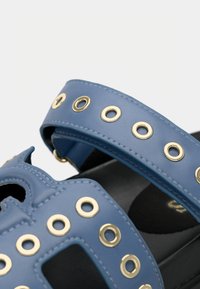 Μπλε συνθετικά σανδάλια με χρυσά μεταλλικά στοιχεία, σχέδιο με κοψίματα και διακοσμητικές τρύπες στα λουριά. Ορατή υφή μαύρης σόλας από κάτω.