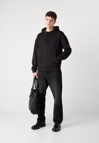 Schwarzer Baumwoll-Hoodie mit Vorderentasche, blauem Logo auf der Brust. Kombiniert mit dunklen Jeans und schwarzen Schuhen, hält eine strukturierte schwarze Tragetasche.