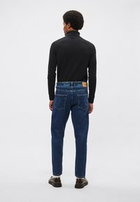 Blue denim jeans with a classic fit, featuring a leather patch at the waist, and a smooth texture. Worn with a black turtleneck sweater.