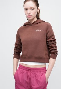 Žena u smešnom Nike dukserici smeđe boje i ružičastim hlačama sa podešavanjem, stoji sa rukama u džepovima na jednostavnoj svetloj pozadini.