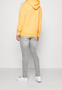 Ljusgul hoodie, grå smala jeans, vita skor med gröna detaljer. Hoodien har dragsko, ribbade muddar och en strukturerad yta.