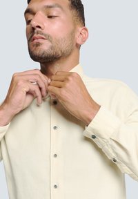 Lichtgele button-up shirt met een bandkraag, zachte textuur, knoopsluitingen en lange mouwen. Model past de kraag met de handen aan.