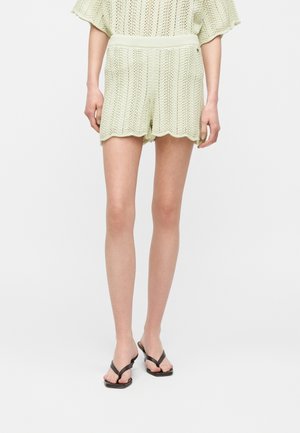 Lichtgroene gebreide korte broek met een geschulpte zoom, gedragen met een bijpassend topje en zwarte sandalen met open teen op een witte achtergrond.