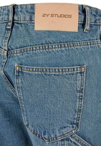 Blugii din denim albastru deschis au o etichetă din piele cu inscripția „2Y STUDIOS” în relief, cusături contrastante și un buzunar în spate pentru funcționalitate.