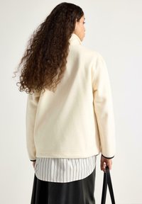 Žena s dlouhými kudrnatými vlasy, oblečená v krémové fleecové bundě, pruhovaném tričku, černé sukni, držící černou kabelku, otočená zády na bílém pozadí.