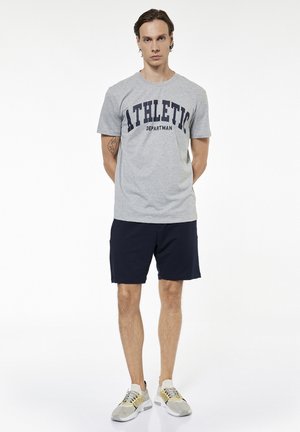 Man staat met handen achter de rug, draagt een grijs T-shirt met "ATHLETIC DEPARTMAN", zwarte shorts en beige-witte sneakers op een witte achtergrond.