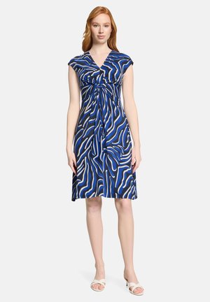 Femme portant une robe mi-longue à motif abstrait bleu, noir et blanc, avec des manches courtes et un détail noué à la taille, debout en sandales blanches.