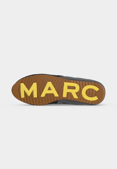 Semelle de chaussure avec bande de roulement marron et grandes lettres jaunes formant "MARC" au bas, sur un fond uni.