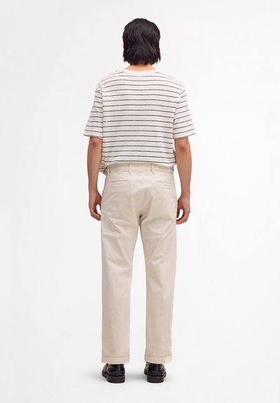 Personne aux cheveux courts et foncés portant une chemise blanche à rayures à manches courtes, un pantalon beige et des chaussures noires, debout de dos sur un fond blanc.