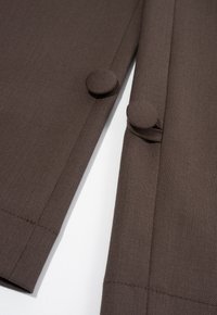 Bruine stoffen kledingdetail met twee ronde knopen en een gestructureerde weving. Gestikte zoom met een gladde afwerking.