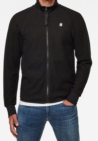 Svart zip-up jacka med hög krage, ribbade manschetter och sidofickor. Har en liten logotyp på vänster bröst och en jämn textur.